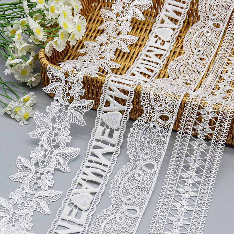 Assorted white water soluble lace trims featuring intricate floral, script, and geometric embroidery patterns displayed on a woven basket.