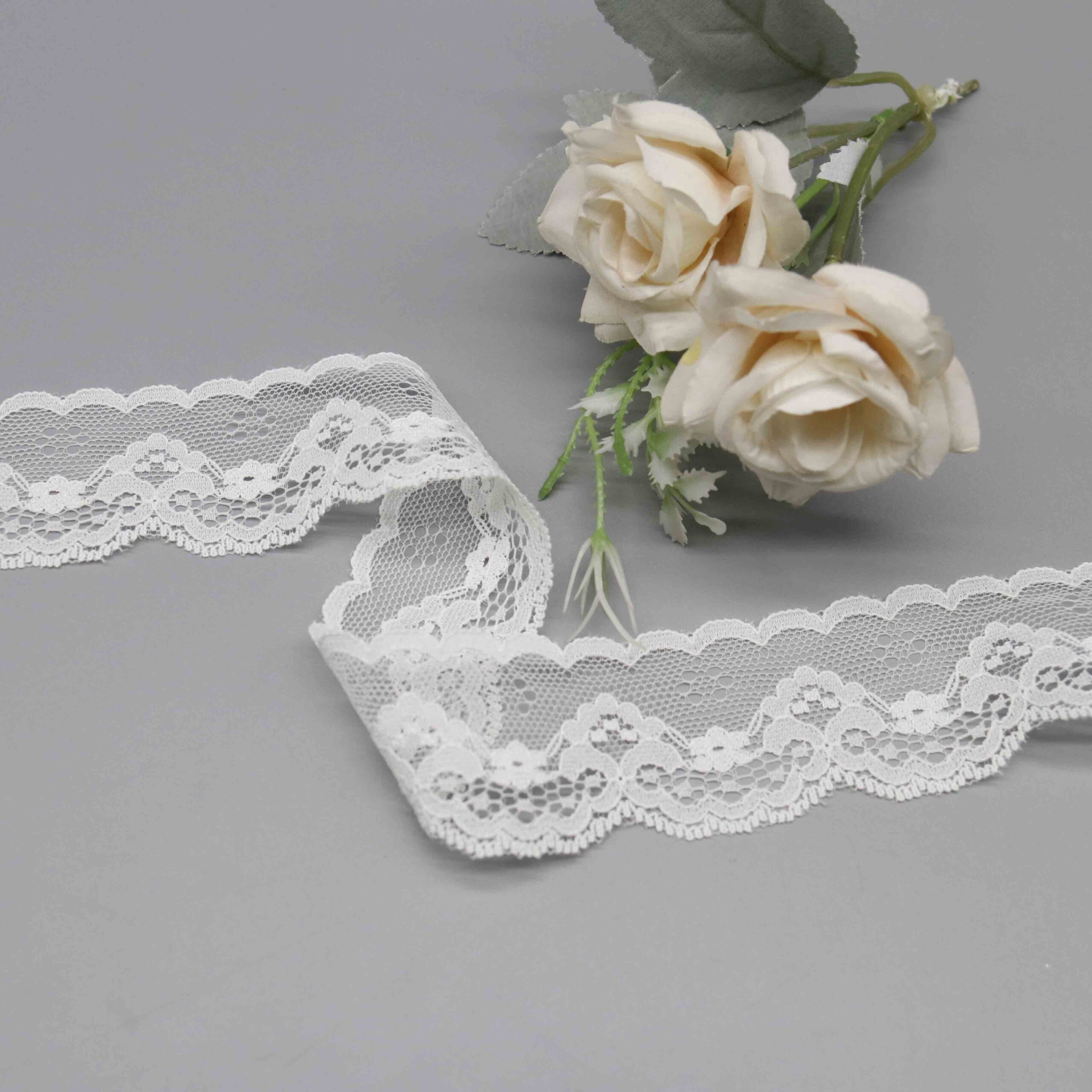 High-resolution image of pure white lace trim ribbon featuring a fine nylon mesh foundation, asymmetric design with a sew-friendly straight side and flowing scalloped arcs blooming with detailed floral embroidery; airy texture contrasts the stitched flowers, perfect for bridal dress cuffs and elegant garment accents.