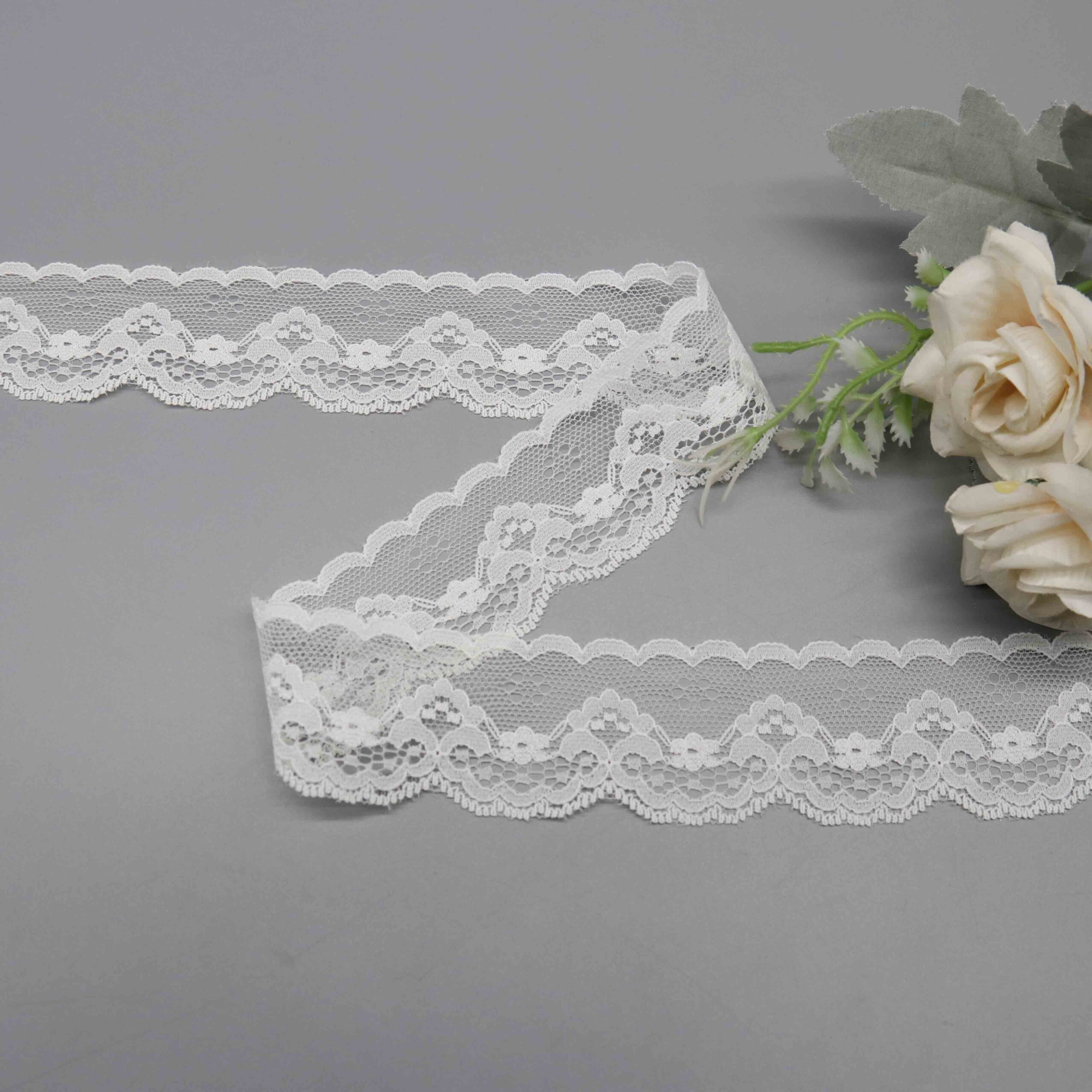 Sophisticated display of pure white lace trim ribbon with a breathable nylon mesh base, asymmetric structure including a straight edge for attachment and scalloped wave side highlighted by full, embroidered flower designs; smooth lines and airy feel enhance bridal and dress embellishments beautifully.