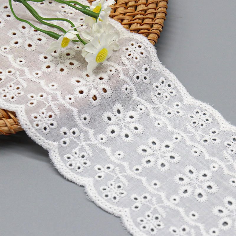 A close-up studio shot of a wide 8cm 100% cotton eyelet lace with a floral medallion pattern and scalloped edges, next to a bouquet of daisies.