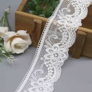 A white floral stretch lace trim with one scalloped edge and one straight edge, shown on a grey background next to a wooden box